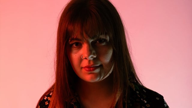 Ellie Spirrett, a woman with a full fringe and long brown hair, she is lit with red lighting