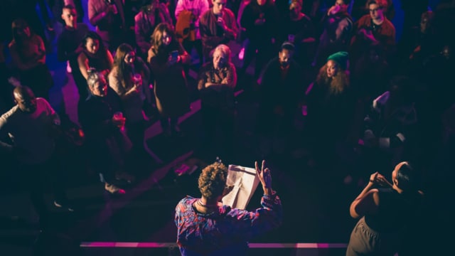 a dimly lit crowd surround a speaker with notes
