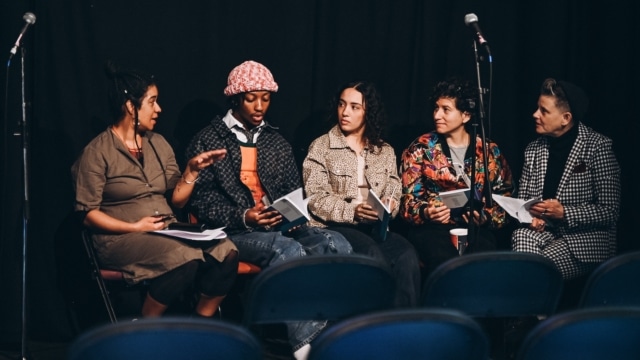 Five writers sit on a stage before the audience has entered. They have books open and are talking.