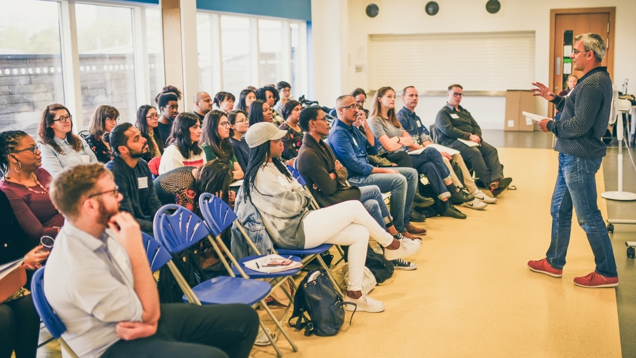 A man stands in front of a crowd of adult students, explaining something with his hands.