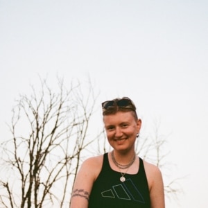 Finn, a white non-binary person in their early thirties, smiles towards the camera. Their hair is pushed back by sunglasses, and they are wearing a black sleeveless top with silver chains around their neck. They are outside and the sky behind them is nearly white, the tree in the background leaf-less.