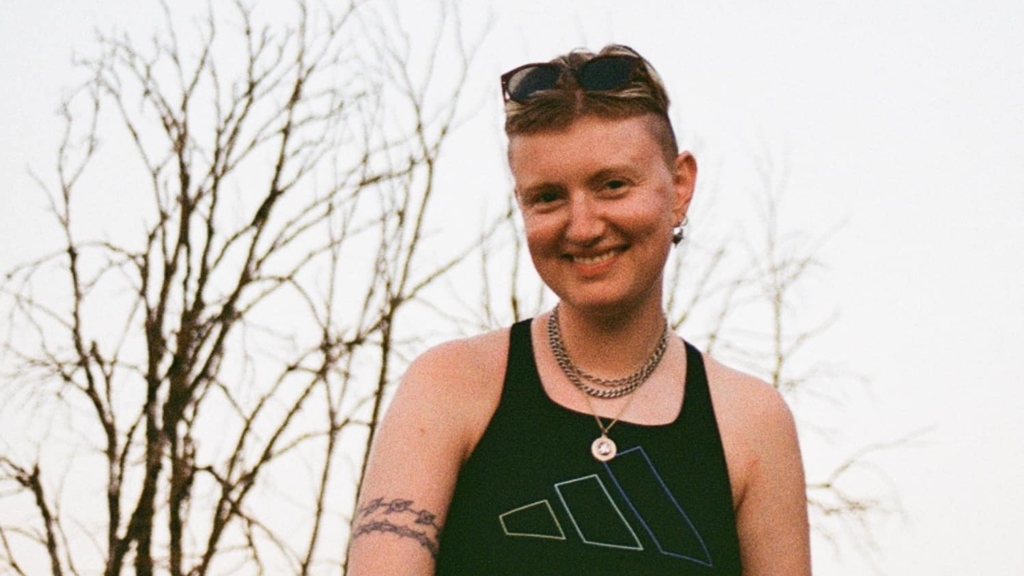 Finn, a white non-binary person in their early thirties, smiles towards the camera. Their hair is pushed back by sunglasses, and they are wearing a black sleeveless top with silver chains around their neck. They are outside and the sky behind them is nearly white, the tree in the background leaf-less.