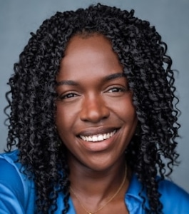 A dark-skinned woman with black curly hair smiles while looking directly at the camera. She wears a blue shirt and a thin, gold necklace with a square, gold pendant. She is sat in a relaxed pose in front of a blue-grey background.