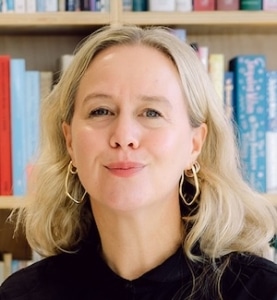 A light-skinned woman with long blond hair is standing against a bookshelf with brightly coloured books and pine shelves. She wears a black jacket and large, gold hoop earrings.